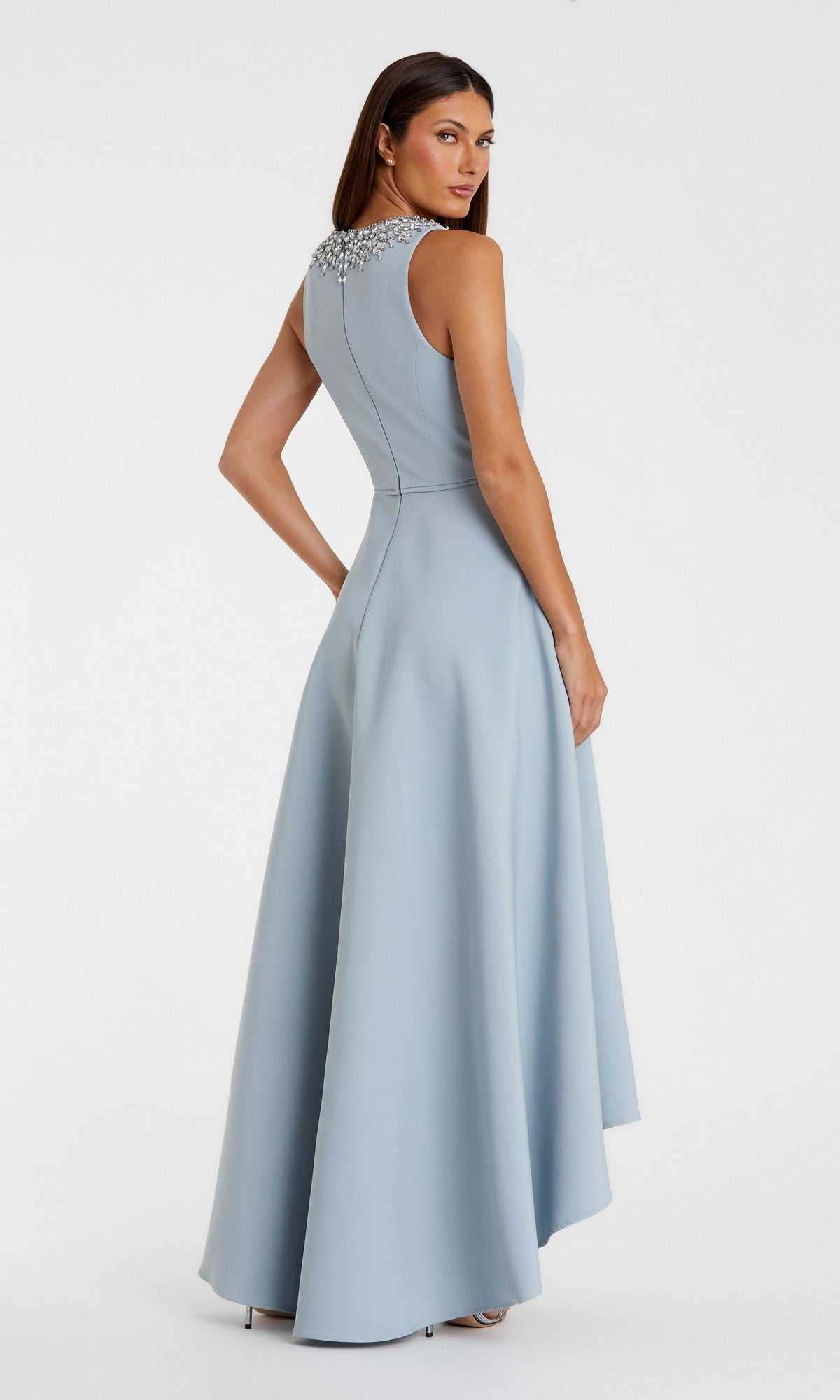 Woman models the Mac Duggal 49844 High-Low Formal Dress, featuring a sleeveless design, embellished neckline, and flowing A-line skirt, standing against a plain background and looking over her shoulder.